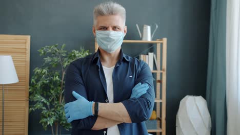 Portrait-of-adult-man-wearing-preventive-medical-mask-and-gloves-standing-in-apartment-with-arms-crossed
