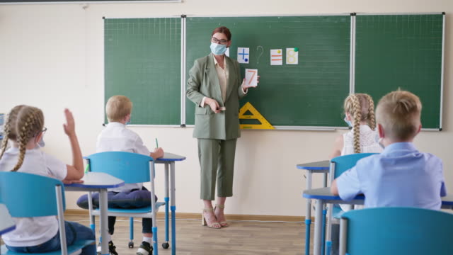 study-during-pandemic,-teacher-in-medical-mask-and-glasses-at-lesson-near-whiteboard-asks-pupils-questions,-schoolgirl-is-ready-to-answer-and-raises-hand