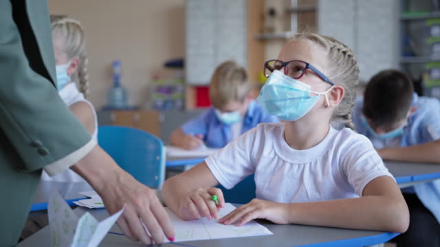 pupils-back-at-school-after-quarantine-and-lockdown,-schoolgirl-in-glasses-and-medical-mask-doing-classwork-sitting-at-desk