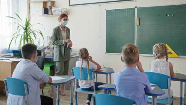 back-to-school,-female-teacher-in-glasses-shows-a-pupils-how-to-put-on-medical-mask-on-face-before-lesson-in-classroom