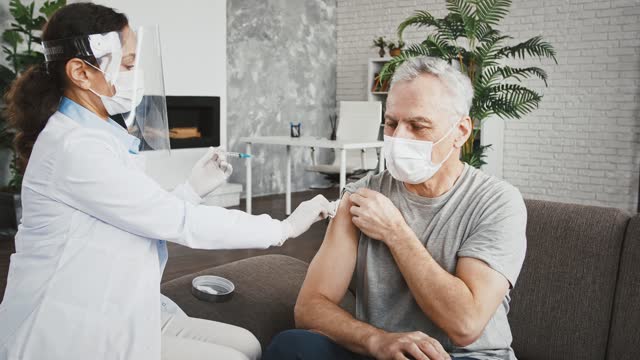 Frau-Arzt-in-Schutzmaske-und-Gesichtsschild-injiziert-Impfstoff-in-die-Schulter-des-alten-Mannes,-der-auf-dem-Sofa-zu-Hause-sitzt