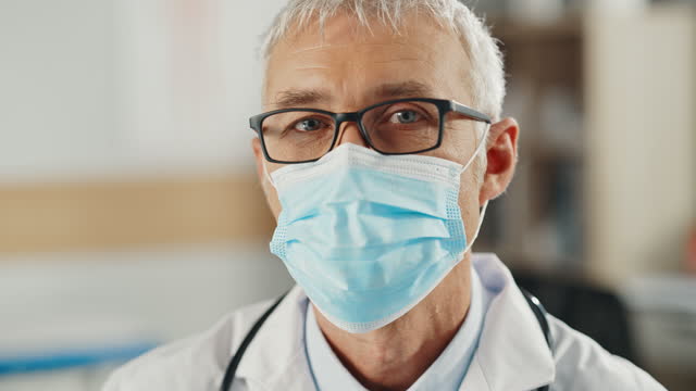 Close-Up-Portrait-of-a-Senior-Caucasian-Male-Doctor-or-Surgeon-Wearing-a-Protective-Face-Mask-and-Glasses.-Middle-Aged-Scientist-Calmly-Looking-at-Camera.-Medical-Care-Specialist-in-Covid-19-Reality.