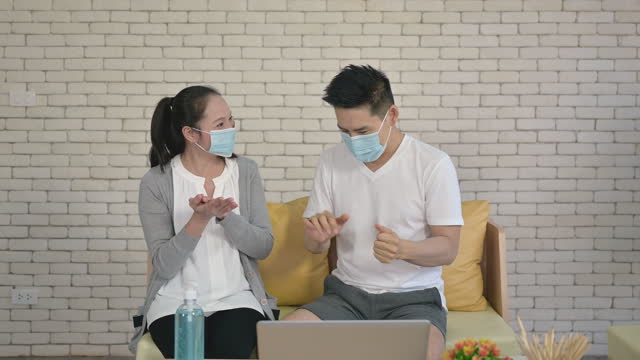 Asiatische-erwachsene-Paar-sitzt-auf-der-Couch-arbeiten-zu-Hause-tragen-Hygiene-Gesichtsmaske-und-waschen-Hände-mit-Alkohol-Gel-oder-Desinfektionsmittel-zu-schützenden-Pandemie-des-Coronavirus-oder-Covid-19.