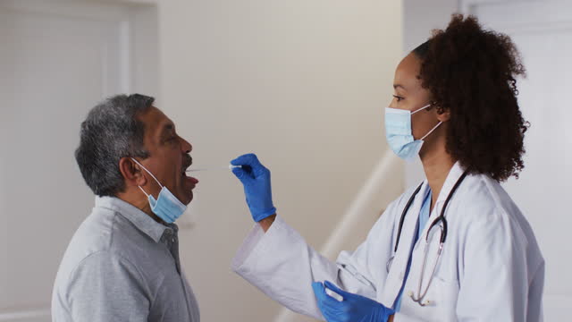 Mixed-race-female-doctor-wearing-mask-doing-swab-test-on-senior-man-at-home