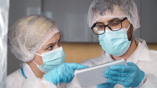 Ärzteteam-in-weißen-Mänteln,-Handschuhen-und-Schutzmasken-diskutiert-Patientendiagnose-auf-digitaler-Tablette-im-Krankenhaus.-Behandlungskonzept-für-Coronavirus,-covid-19