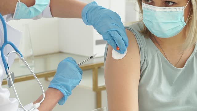 Close-up-of-medical-nurse-in-gloves-and-mask-vaccinates-female-patient-in-clinic.-Doctor-injects-medicine-into-hand-from-syringe-and-applies-medical-patch.-Covid-19,-coronavirus-vaccine