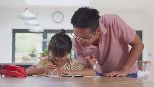 Asiatische-Vater-hilft-Zu-Hause-Schulende-Tochter-arbeiten-am-Tisch-in-Küche-Schreiben-in-Buch
