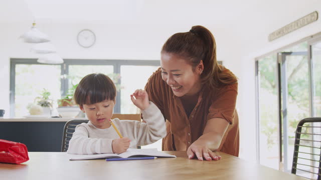 Madre-asiática-ayudando-a-su-hijo-escolar-en-casa-trabajando-en-la-mesa-en-la-cocina-escribiendo-en-el-libro