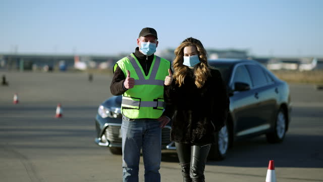 Confident-Caucasian-woman-and-man-in-Covid-face-masks-showing-thumbs-up-with-car-at-background.-Portrait-of-driving-instructor-and-student-posing-on-coronavirus-pandemic-outdoors
