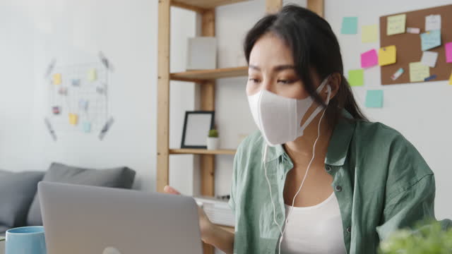 Asiatische-Geschäftsfrau-trägt-medizinische-Gesichtsmaske-mit-Laptop-sprechen-mit-Kollegen-über-Plan-in-Videoanruf-während-der-Arbeit-von-zu-Hause-aus-im-Wohnzimmer.