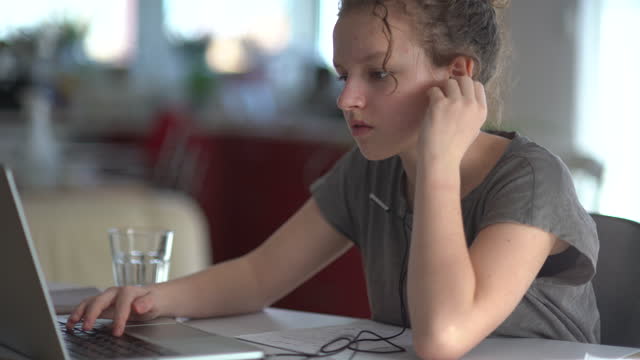 Niños,-educación-y-concepto-de-aprendizaje-distante---adolescente-estudiante-en-auriculares-con-computadora-portátil-en-casa.-Linda-chica-escucha-atentamente,-responde-y-escribe-en-un-cuaderno