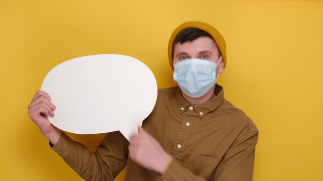 Portrait-of-amazed-young-man-in-medical-protective-face-mask-holding-and-pointing-at-blank-speech-bubble,-wears-shirt-and-beanie-hat,-isolated-on-yellow-studio-background.-People-lifestyle-concept