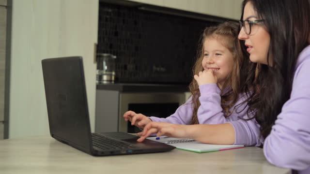 Mom-and-daughter-are-sitting-at-a-table-in-the-kitchen.-Mom-teaches-her-little-daughter-to-use-a-laptop-and-the-Internet.