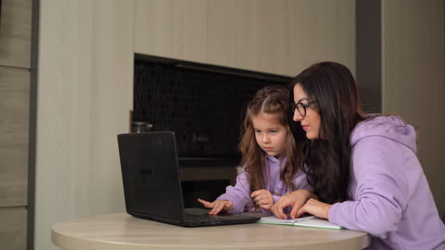 Mamá-e-hija-están-sentadas-juntas-en-casa-en-la-cocina-de-la-mesa.-Una-mujer-enseña-a-un-niño-a-usar-Internet-y-una-computadora-portátil.