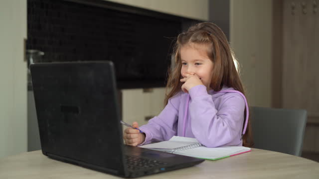 Fernunterrichtskonzept.-eine-kleine-Schülerin-studiert-online-mit-einem-Laptop,-Notizen-in-einem-Notizbuch-machen.-das-Kind-sitzt-am-Tisch