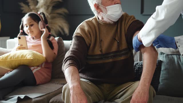 Female-doctor-visiting-senior--man-giving-her-vaccination.-both-wearing-face-masks.-retirement-health-care-at-home,--Grandfather-and-granddaughter-in-isolation.-Vaccination-of-pensioners-at-home
