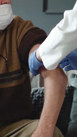 Female-doctor-visiting-senior--man-giving-her-vaccination.-both-wearing-face-masks.-retirement-health-care-at-home,--Grandfather-and-granddaughter-in-isolation.-Vaccination-of-pensioners-at-home.-Vertical-screen