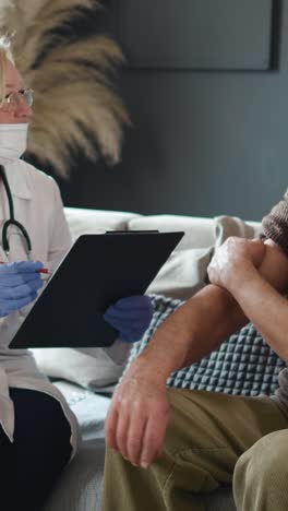 Doctor-advises-and-gives-therapy-to-the-old-grandfather-after-the-vaccination-against-COVID-19.-Vaccination-people-at-home.--Girl-child--with-the-headphones-uses-a-mobile-phone.-Vertical-screen