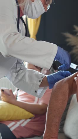 Vertical-screen.-Female-doctor-gives-the-vaccine-to-senior-man-against-COVID-19,-the-little-girl-uses-a-mobile-phone-in-the-background.-Family-health,-doctor-home-visit