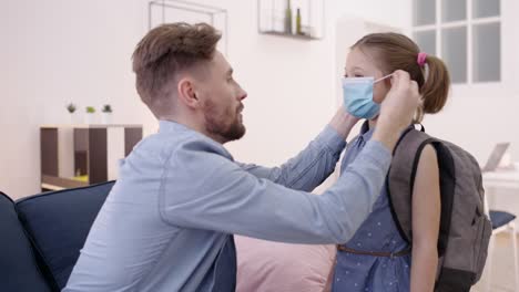 Padre-cariñoso-preparando-a-su-hija-a-la-escuela-durante-el-coronavirus,-poniéndose-la-máscara