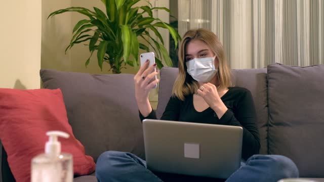 Young-Turkish-woman-with-a-facial-mask,-having-a-video-call-on-her-smartphone-while-sitting-on-the-sofa.-There-is-a-laptop-computer-on-her-lap,-coffee-cup-and-a-hand-sanitizer-bottole-on-the-table.