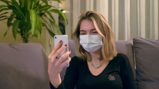 Young-Turkish-woman-with-a-facial-mask,-having-a-video-call-on-her-smartphone-while-sitting-on-the-sofa.-There-is-a-laptop-computer-on-her-lap,-coffee-cup-and-a-hand-sanitizer-bottole-on-the-table.