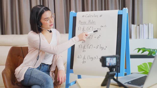 Asian-school-young-woman-teacher-working-from-home-teach-online-math-subject-to-student-studying-from-home.-Girl-points-on-whiteboard,-talk-on-headphone.-Remote-education-class-during-covid19-pandemic