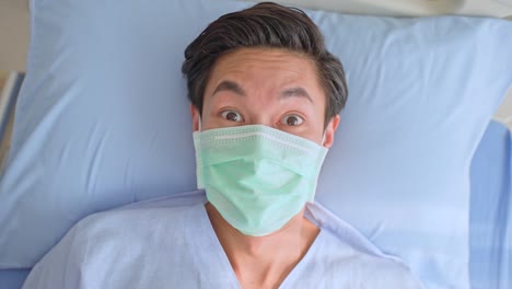 Asiatische-junge-männliche-Patient-trägt-schützende-Gesichtsmaske-auf-dem-Bett-liegen,-warten-auf-die-Behandlung-durch-den-Arzt-im-Erholungsraum.-Der-Mann-wacht-erschrocken-und-erschreckt-auf,-fühlt-sich-in-Panik-oder-nervös-von-schlechtem-Traum