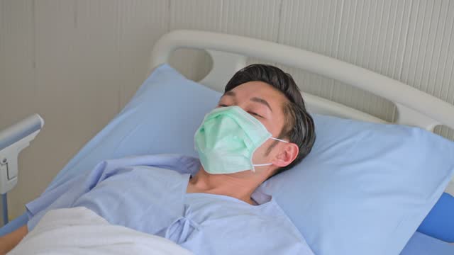 Unwell-young-Asian-male-patient-coughing-while-rest-lying-in-hospital-bed-recovery-after-fell-ill-with-cold-and-flu.-Wearing-face-mask-in-quarantine-for-protection-during-covid-19-wait-for-treatment.