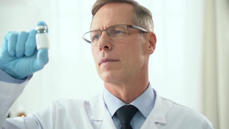 Development-of-the-vaccine.-Close-up-of-middle-aged-male-doctor-holding-and-looking-at-a-glass-bottle-of-vaccine,-standing-in-a-clinic.-Technician-preparing-the-vaccine