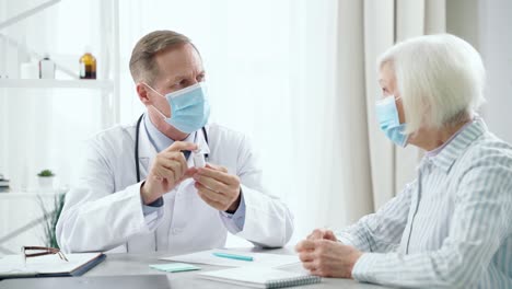 Evite-enfermarse.-Médico-masculino-de-mediana-edad-que-usa-máscara-consultando-a-la-mujer-paciente-de-edad-avanzada,-sosteniendo-y-mostrando-una-botella-de-vidrio-de-la-vacuna,-sentado-en-la-mesa-en-su-oficina