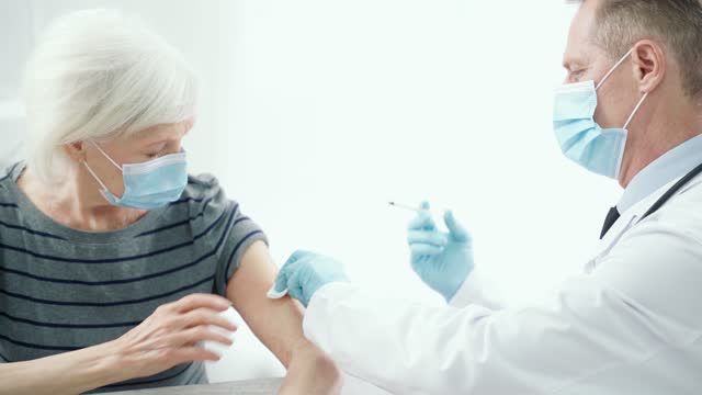 Vacunarse.-Médico-masculino-con-máscara-médica-y-guantes-dando-una-inyección-a-una-anciana-en-el-hospital