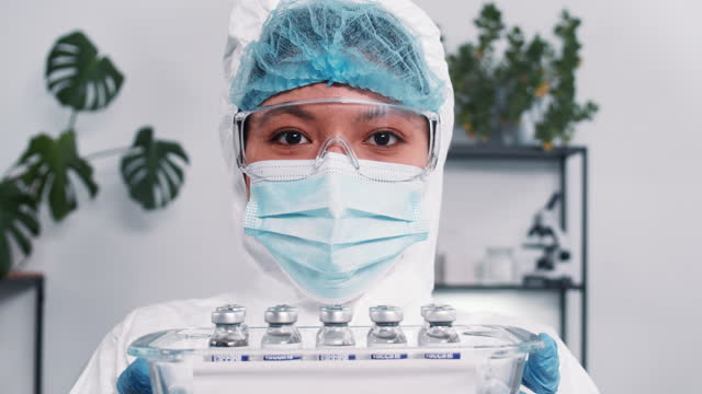 Hoffnung-im-Kampf-gegen-Coronavirus.-Seriöse-multiethnische-Laborwissenschaftlerin-im-Schutzanzug,-die-Tablett-mit-Impfstoffkolben-zeigt.