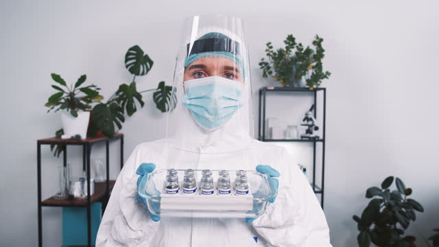 Vaccine-research.-Serious-female-lab-scientist-in-protection-suit-and-shield-holding-cure-flasks-against-coronavirus.