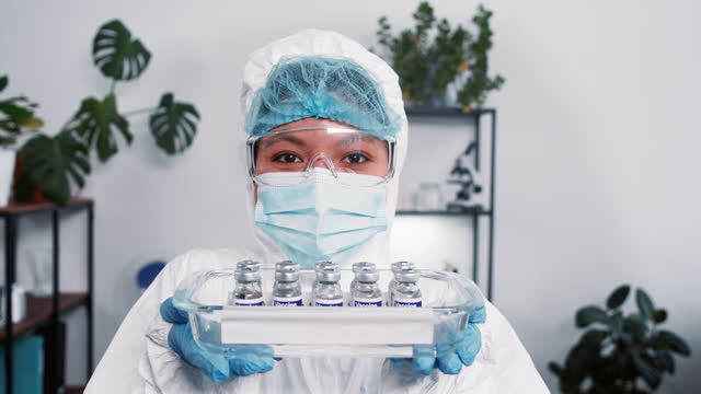 Victory-over-coronavirus.-Cheerful-female-science-lab-worker-in-protection-suit-shows-medical-tray-with-vaccine-flasks.