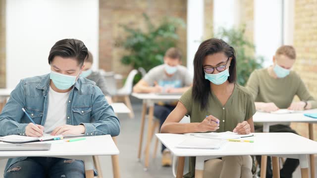 Enthusiastic-learner.-Diverse-students-sitting-at-table-in-university,-wearing-protective-face-mask,-listening-and-making-notes.-Guy-raised-his-hand-during-lecture-in-a-classroom