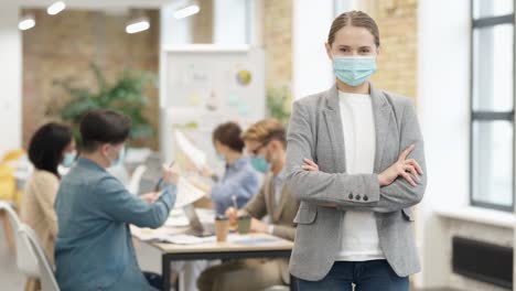 Attractive-caucasian-young-woman-wearing-protective-face-mask-looking-at-camera,-standing-with-arms-crossed-in-a-classroom.-Group-of-diverse-students-working-in-the-background