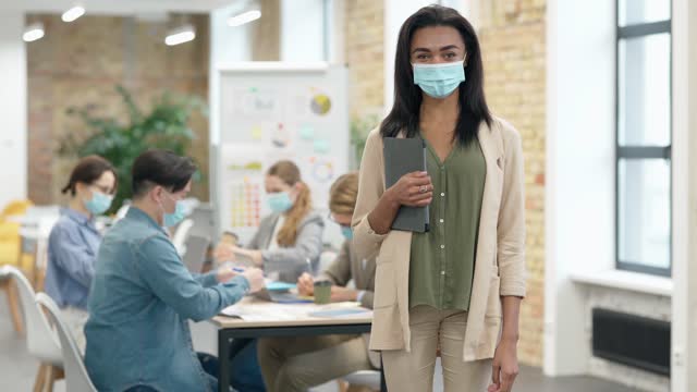 Attractive-mixed-race-young-woman-wearing-protective-face-mask-looking-at-camera,-standing-with-tablet-pc-in-a-classroom.-Group-of-diverse-students-working-in-the-background