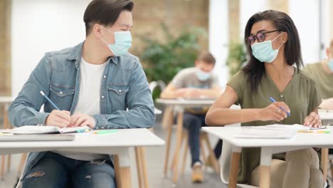 Freunde.-Diverse-Studenten,-die-in-der-Universität-am-Tisch-sitzen,-schützende-Gesichtsmaske-tragen,-Test-schreiben,-betrügen-und-zustimmen,-indem-sie-Ellbogen-in-einem-Klassenzimmer-stoßen