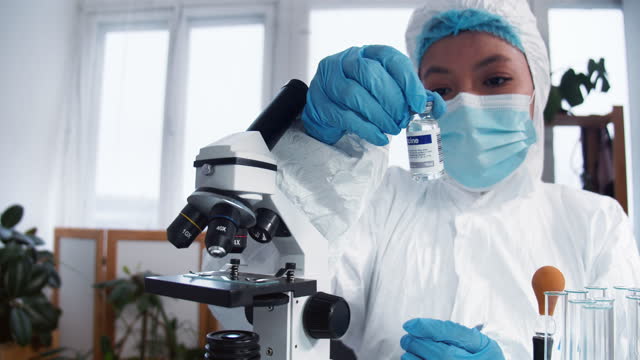 Científica-feliz-del-laboratorio-de-la-clínica-en-traje-de-protección-y-escudo-sosteniendo-frasco-con-nueva-vacuna.-Esperanza-en-la-lucha-contra-las-enfermedades