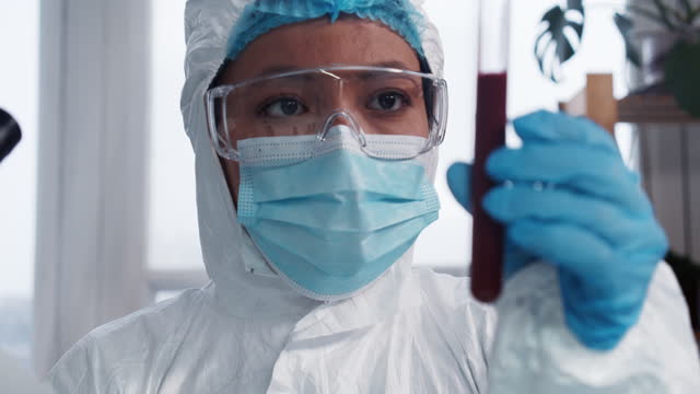 Ensayos-clínicos.-Joven-médico-de-laboratorio-farmacéutico-de-raza-mixta-enfocado-en-traje-de-protección-que-mira-el-tubo-de-ensayo-con-sangre.