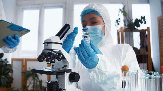 Creating-COVID-19-vaccine.-Two-female-pharmacy-scientists-in-full-protection-suits-test-new-cure-flask-at-clinic-lab.