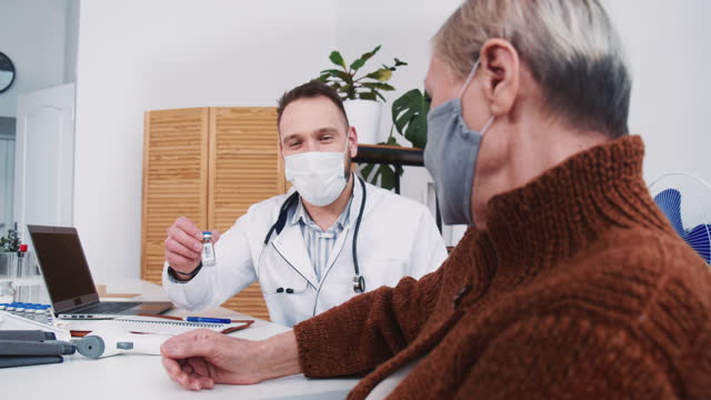 Happy-positive-Caucasian-male-doctor-offers-vaccine-cure-to-senior-grey-haired-patient-woman-in-mask-at-clinic-office.