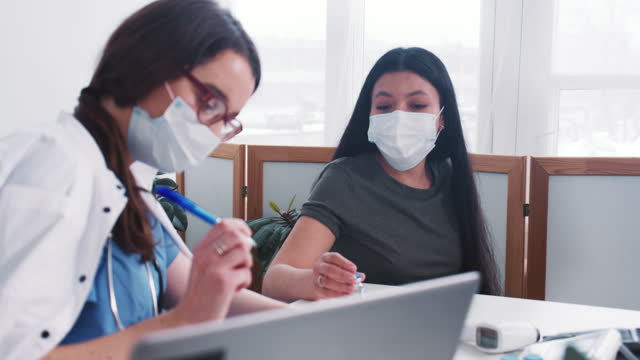 Young-Caucasian-doctor-woman-consults-mixed-race-patient-woman-wearing-mask,-holding-medication-flask-at-medical-exam.