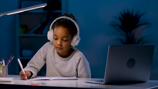 A-girl-in-headphones-takes-notes-while-listening-to-an-audio-lesson,-looks-at-the-laptop,-nods-her-head.-A-girl-sits-at-a-table-under-the-light-of-a-table-lamp.-Close-up.-Slow-motion-ready-59.97fps