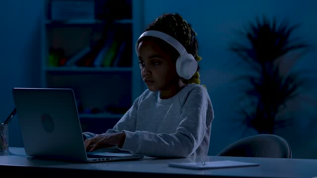 Una-niña-afroamericana-seria-con-grandes-auriculares-blancos-se-sienta-en-una-mesa-en-una-habitación-oscura,-escribiendo-en-su-computadora-portátil.-El-concepto-de-aprendizaje-a-distancia.-De-cerca.-Cámara-lenta-lista-59.97fps