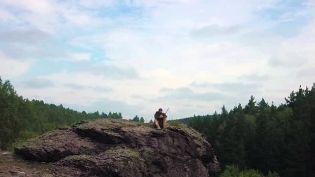 A-sniper-with-a-rifle-sits-on-top-of-the-mountain-and-inspects-the-territory.-View-from-the-drone.-the-camera-moves-from-the-soldier,-revealing-a-view-of-the-landscape