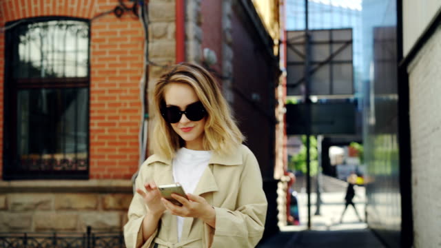 Schöne-junge-Dame-hält-Smartphone-und-benutze-es-zu-Fuß-entlang-der-Straße-in-modernen-Stadt.-Technik,-schöne-glückliche-Menschen-und-Jugend-Lifestyle-Konzept.