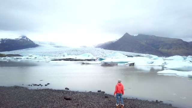 DRONE-aerial-view-of-woman-contemplating-glacier-lagoon-in-Iceland-standing-on-lake-shore