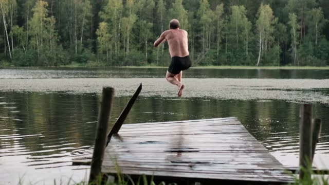 Caucasian-mature-man-jumping-from-wooden-pier-in-lake.